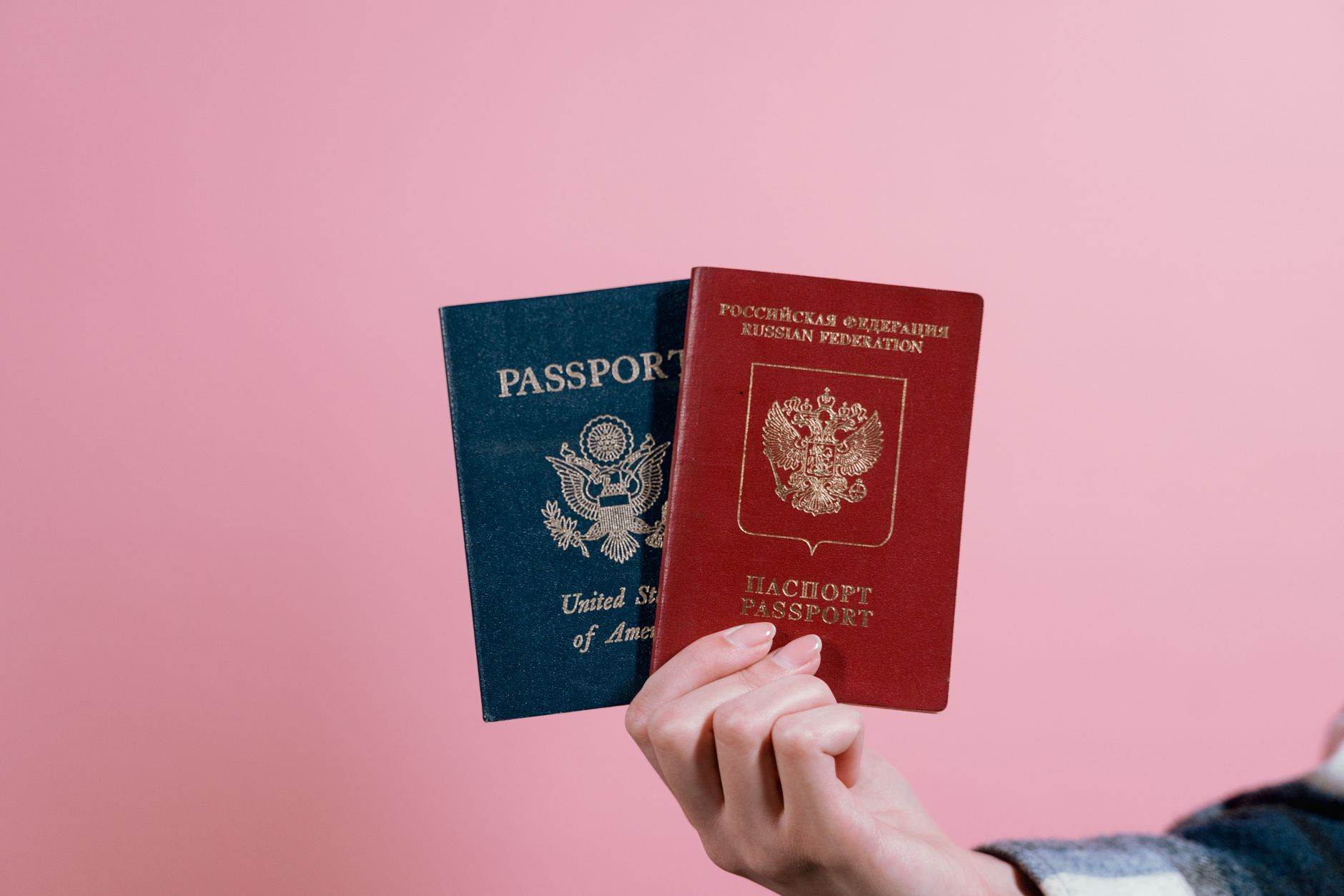 Närbild av en hand som håller ett amerikanskt och ett ryskt pass mot rosa bakgrund.