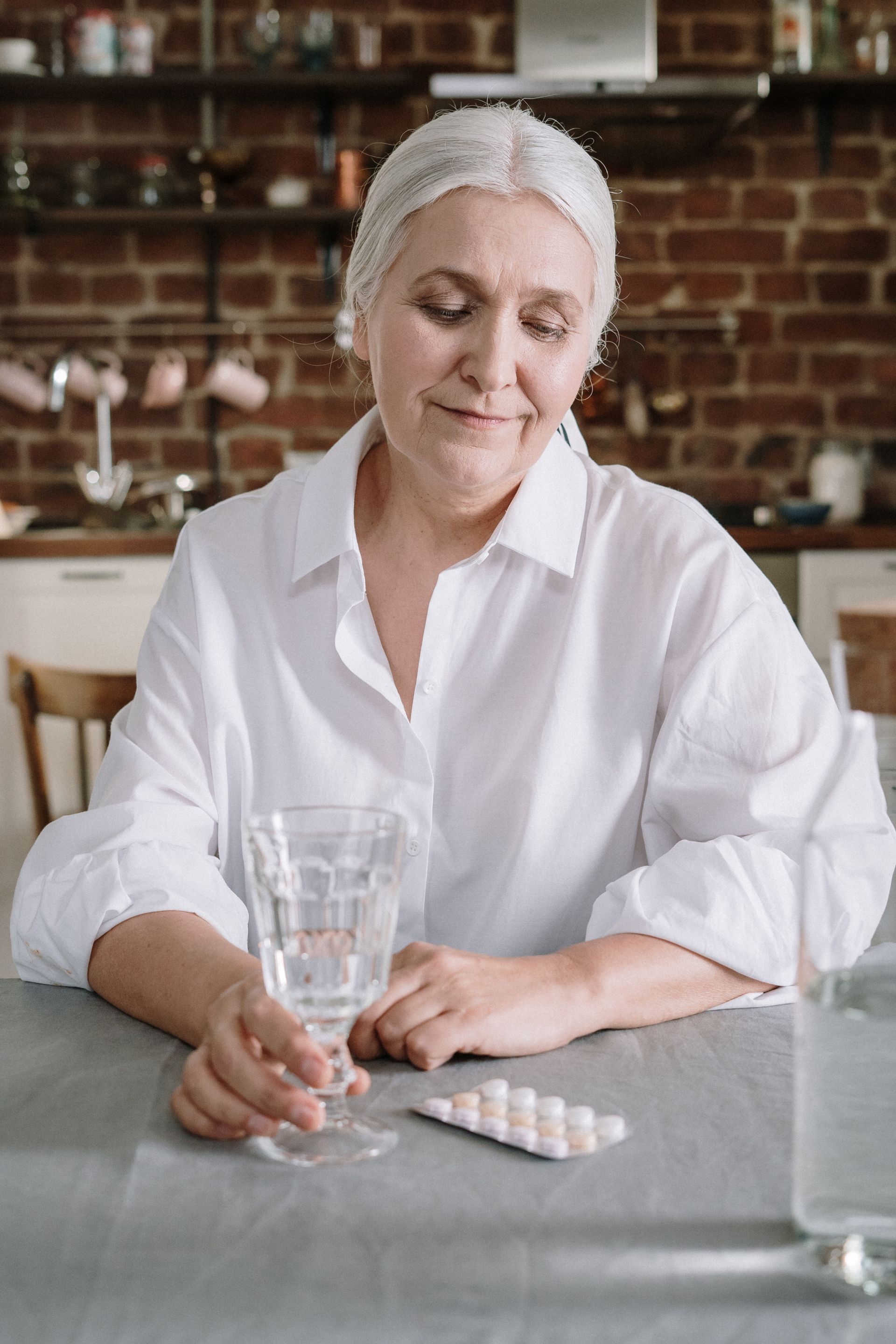 Äldre kvinna sitter vid ett bord hemma med ett glas vatten och mediciner framför sig.