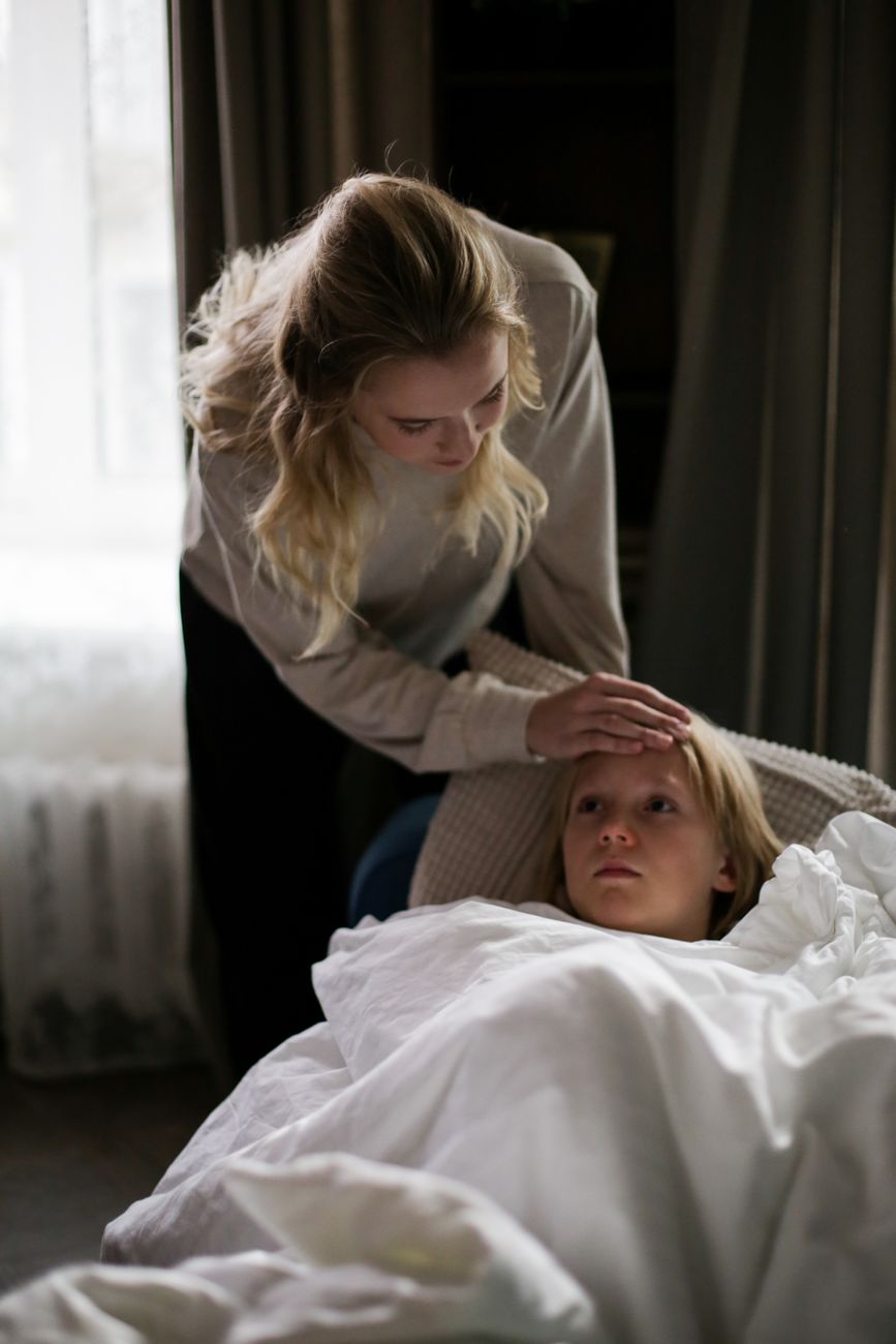 En kvinna sitter hos ett barn som ligger i en soffa under en vit filt.